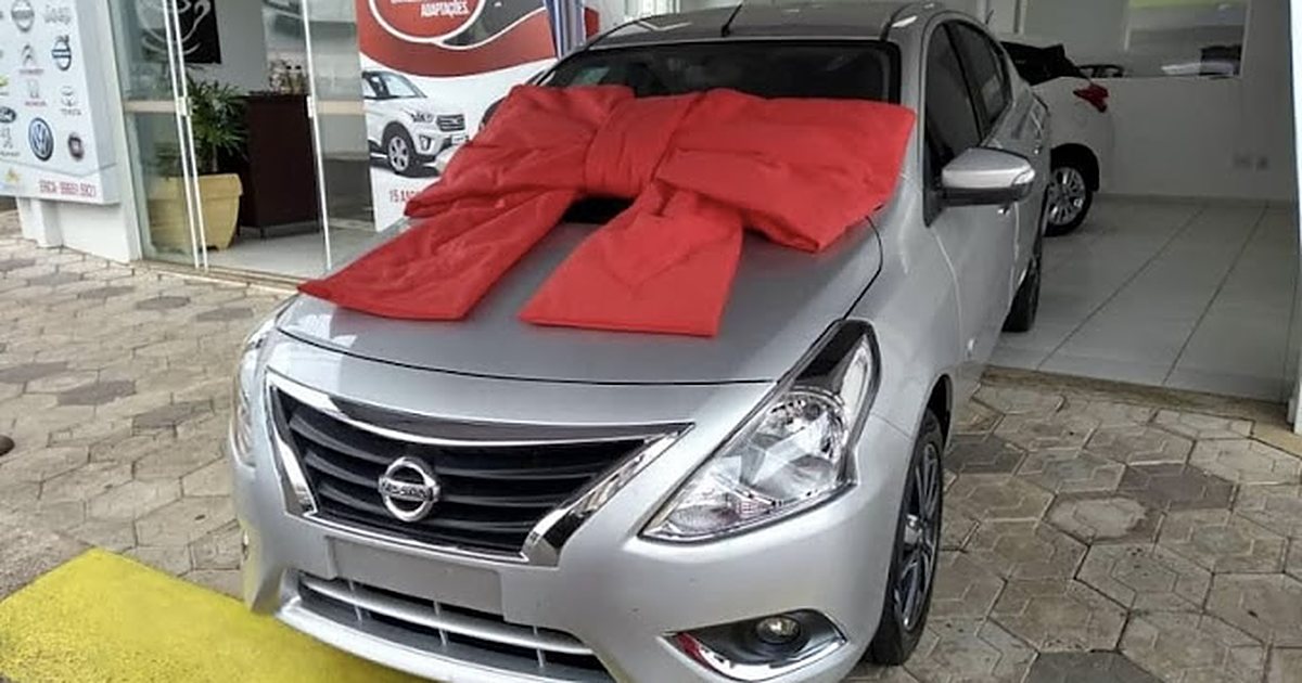 Carro 0km prateado com laço vermelho no pátio de uma concessionária